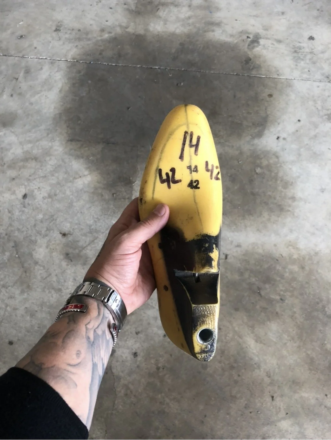 Zapato de seguridad amarillo con marcas de tinta negras, sustentado por una mano con tatuajes y reloj, sobre un suelo de concreto.