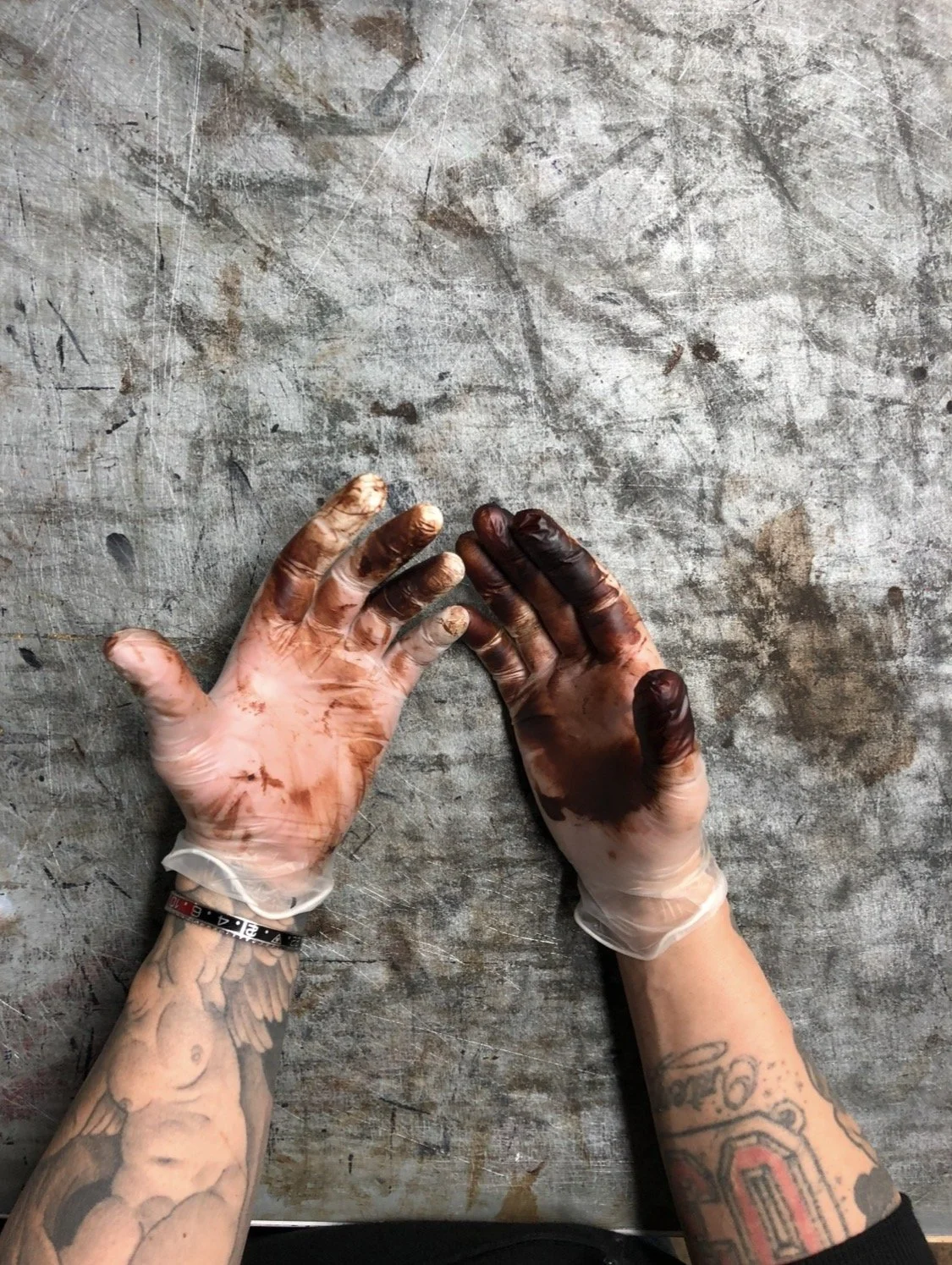 Manos con tinta de tattoo en los dedos y muñecas, con guantes de látex, sobre superficie de mesa de trabajo manchada de tinta y pigmentos.