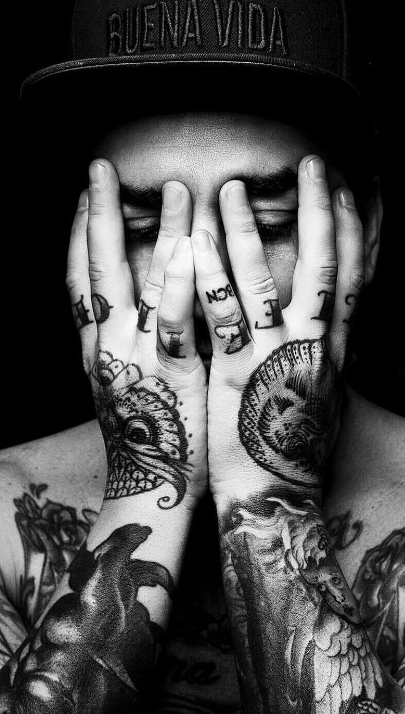 Joven con tatuajes, cubriéndose la cara con las manos, usando gorra con la frase 'Buena Vida' en letras mayúsculas, en blanco y negro.