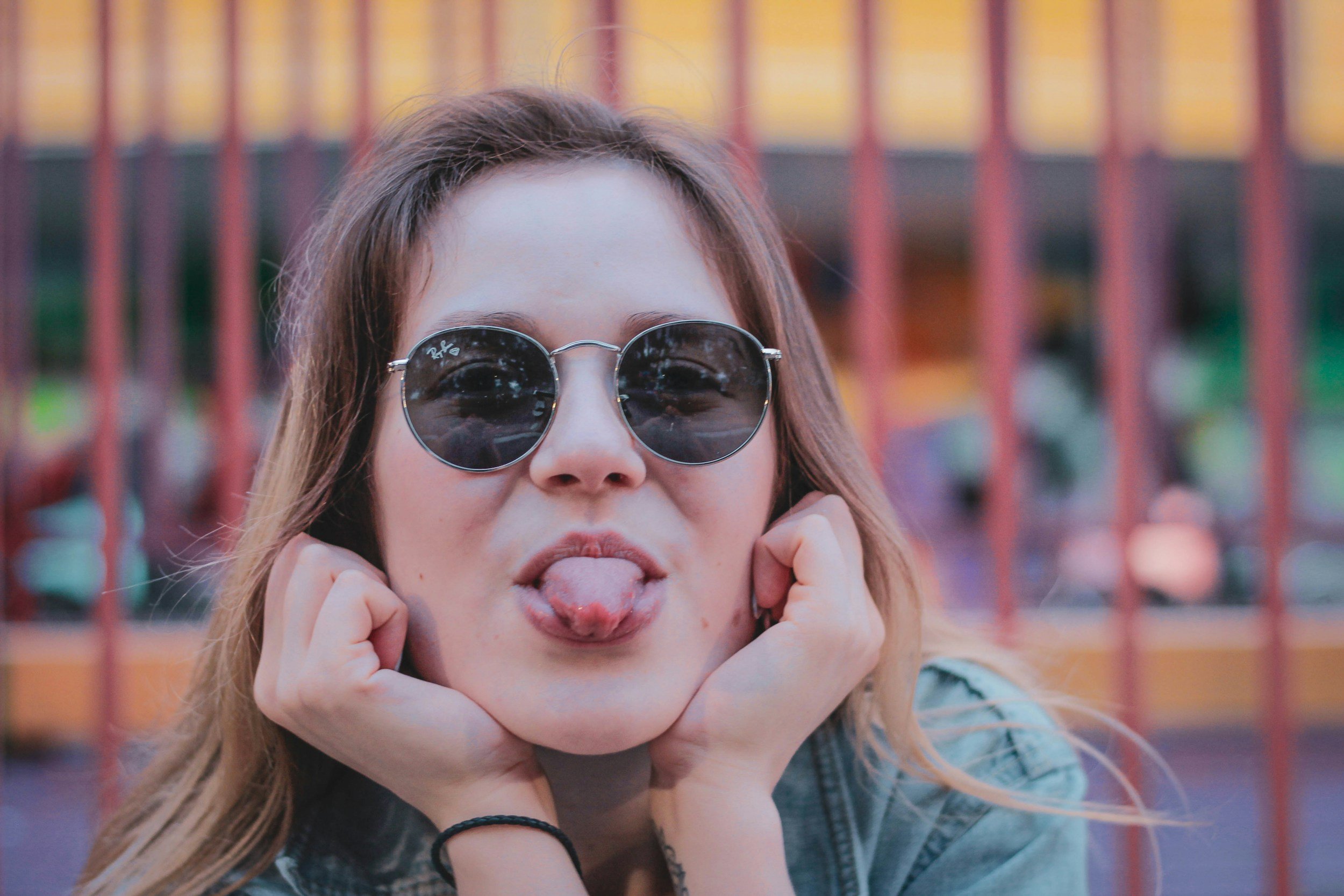 A young woman with long hair wearing sunglasses sticking out her tongue and holding her face with both hands.