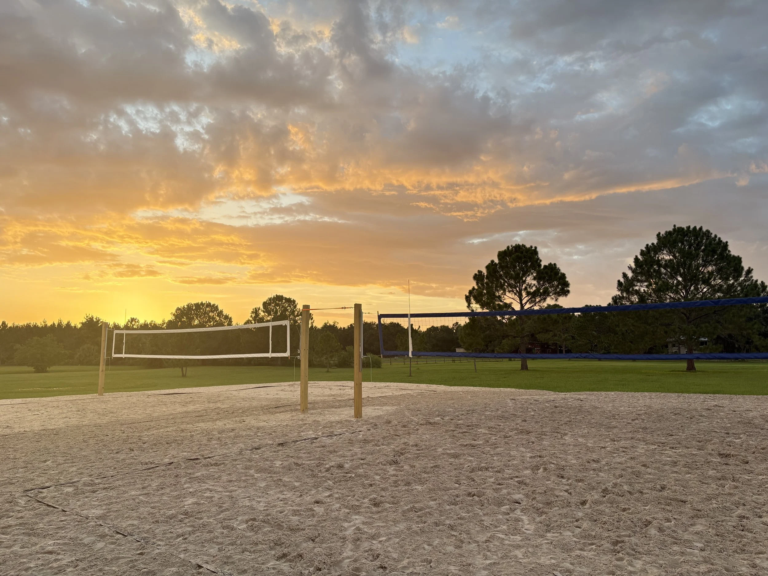 A volleyball net set up on a sandy court outdoors during sunset, with a sky full of clouds and orange hues, and trees in the background.