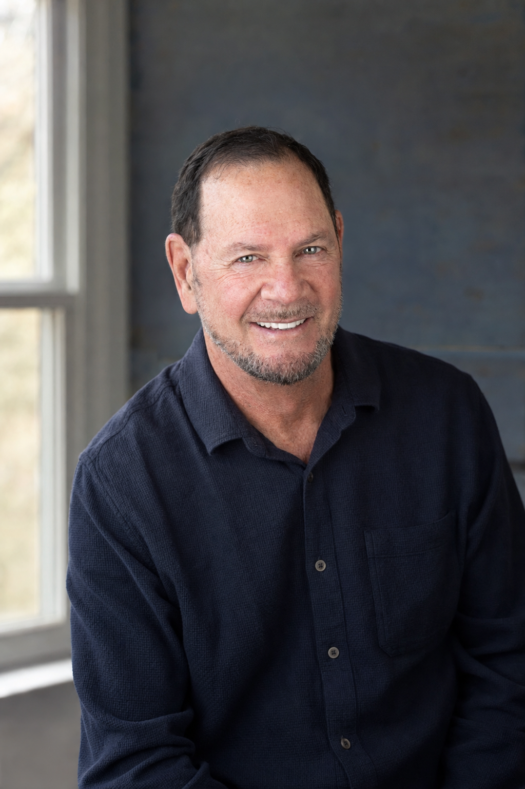 Author Jack Dell smiling beside a window, wearing a dark blue button-down shirt.