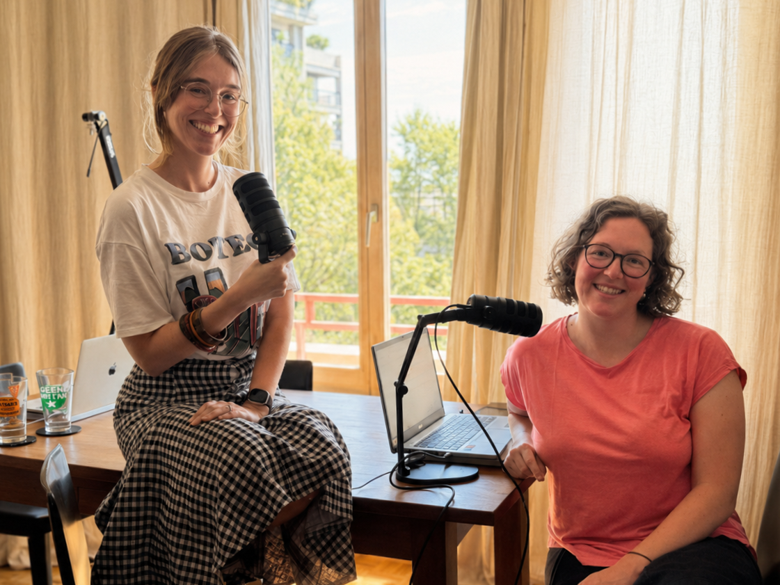 Two women are sitting at a wooden table with microphones and laptops, smiling in a room with large windows and beige curtains, outdoors greenery in background.