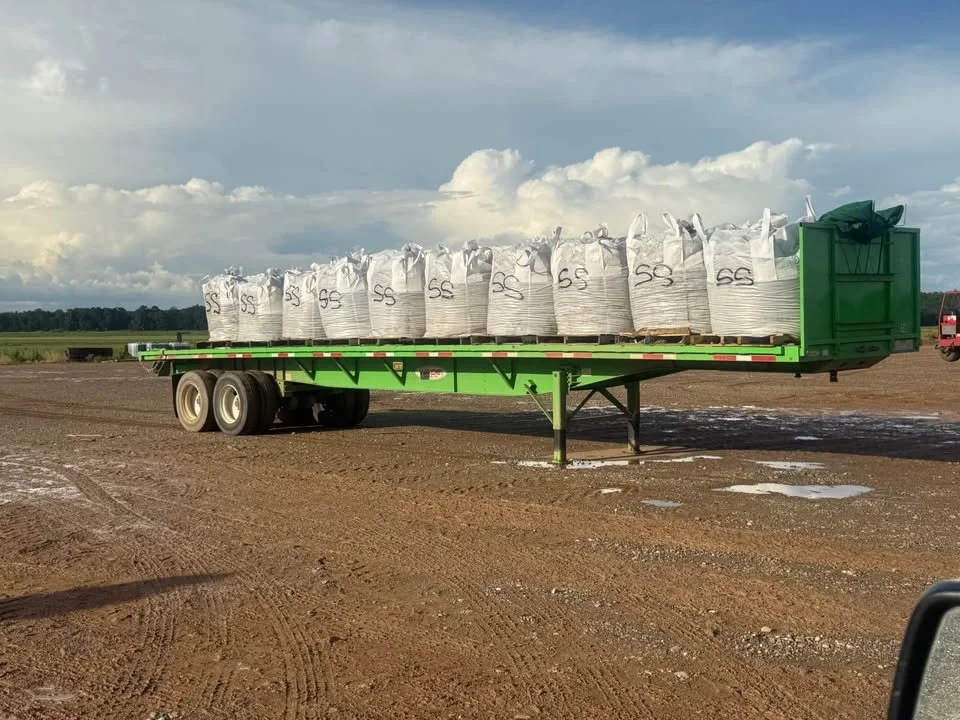 A green flatbed trailer carrying large white bags filled with soil or fertilizer on a dirt field.