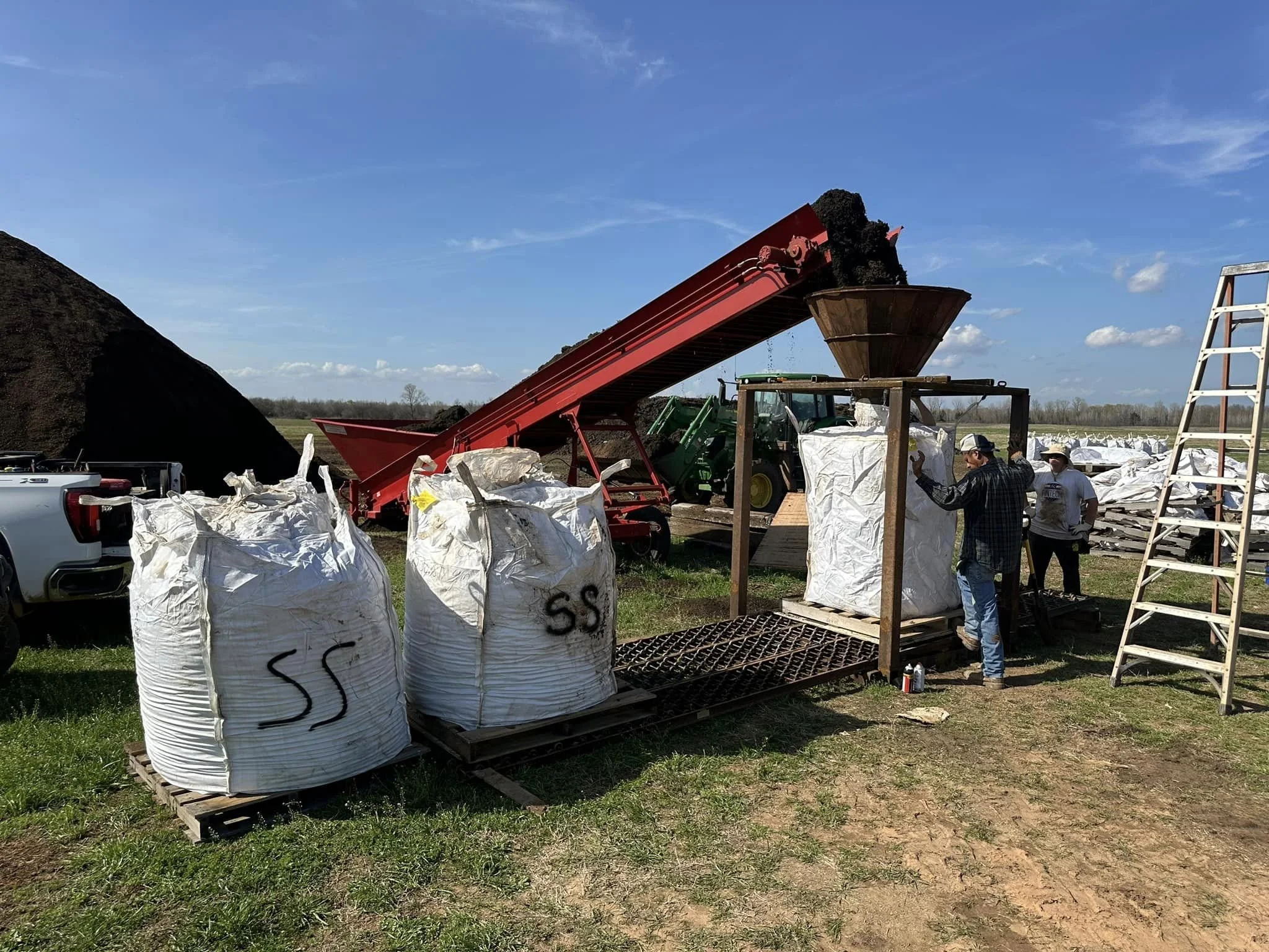 People working outdoors with large white bags, a red conveyor belt moving soil into a large hopper, and trees in the background.