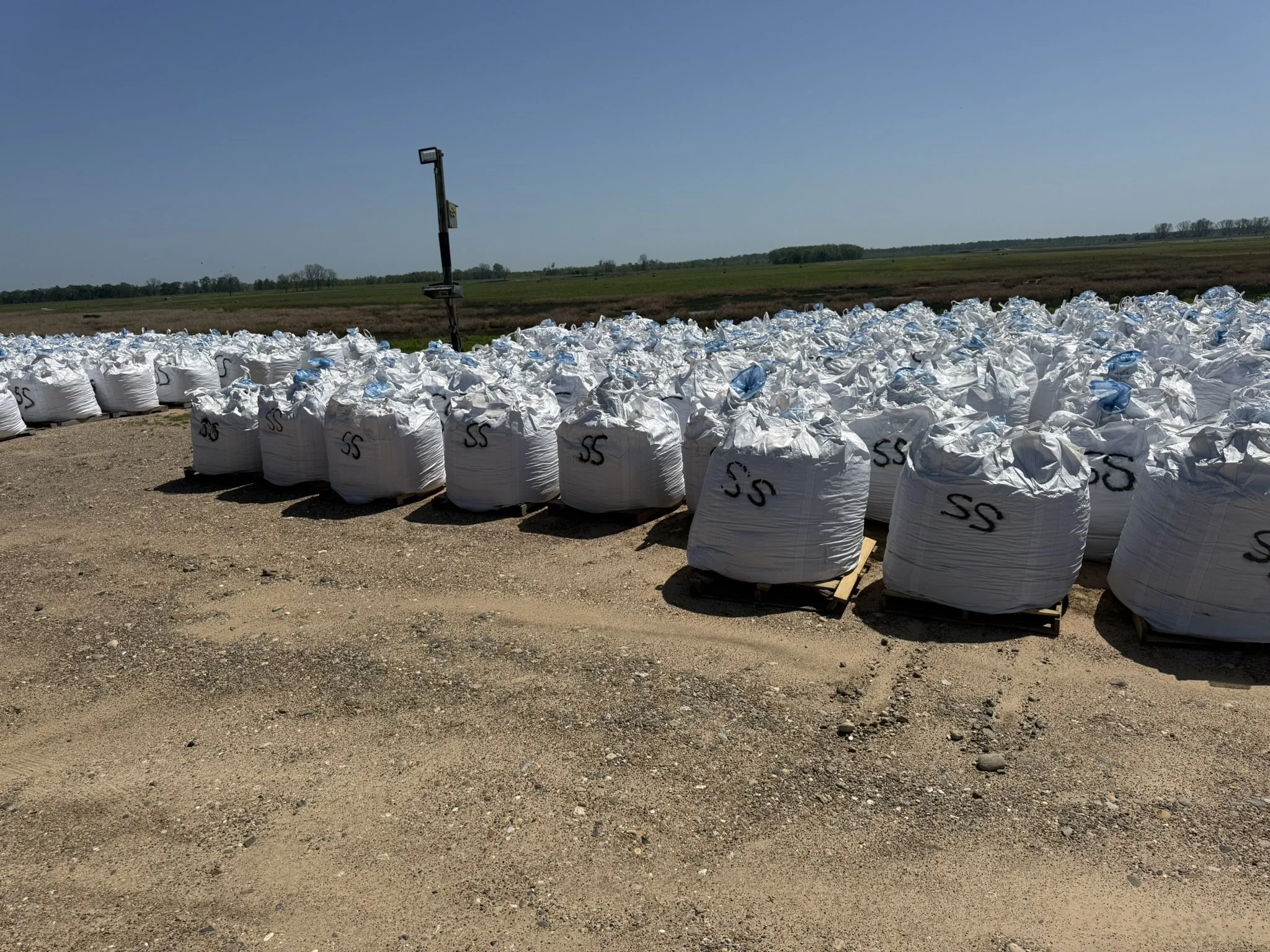 Large white bags of materials with black markings stacked outdoors on a dirt area, with a wide open field and blue sky in the background.