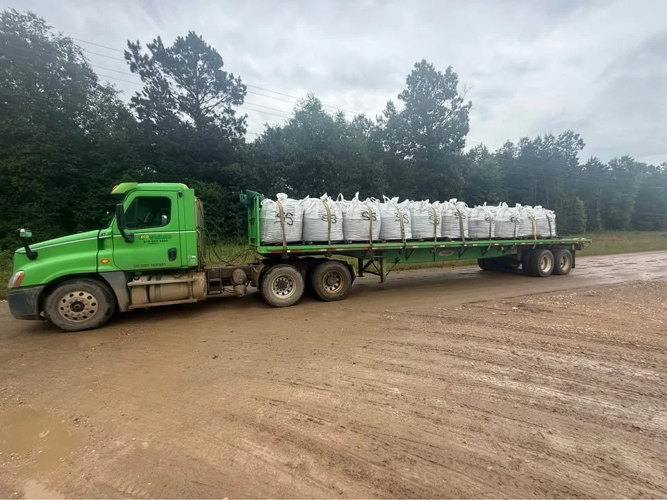A green flatbed truck loaded with large white bags, parked on a dirt road near a wooded area under a cloudy sky.