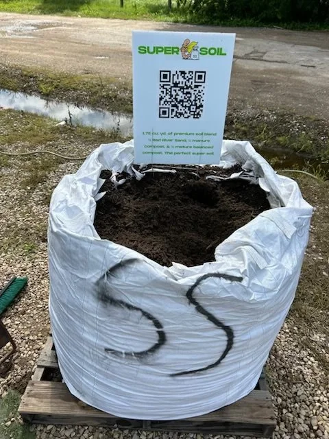 A large white bag filled with dark soil, placed on a wooden pallet outdoors. It has black spray-painted letters 'SS' on it, with a sign behind that includes a QR code and the logo for Super Soil.