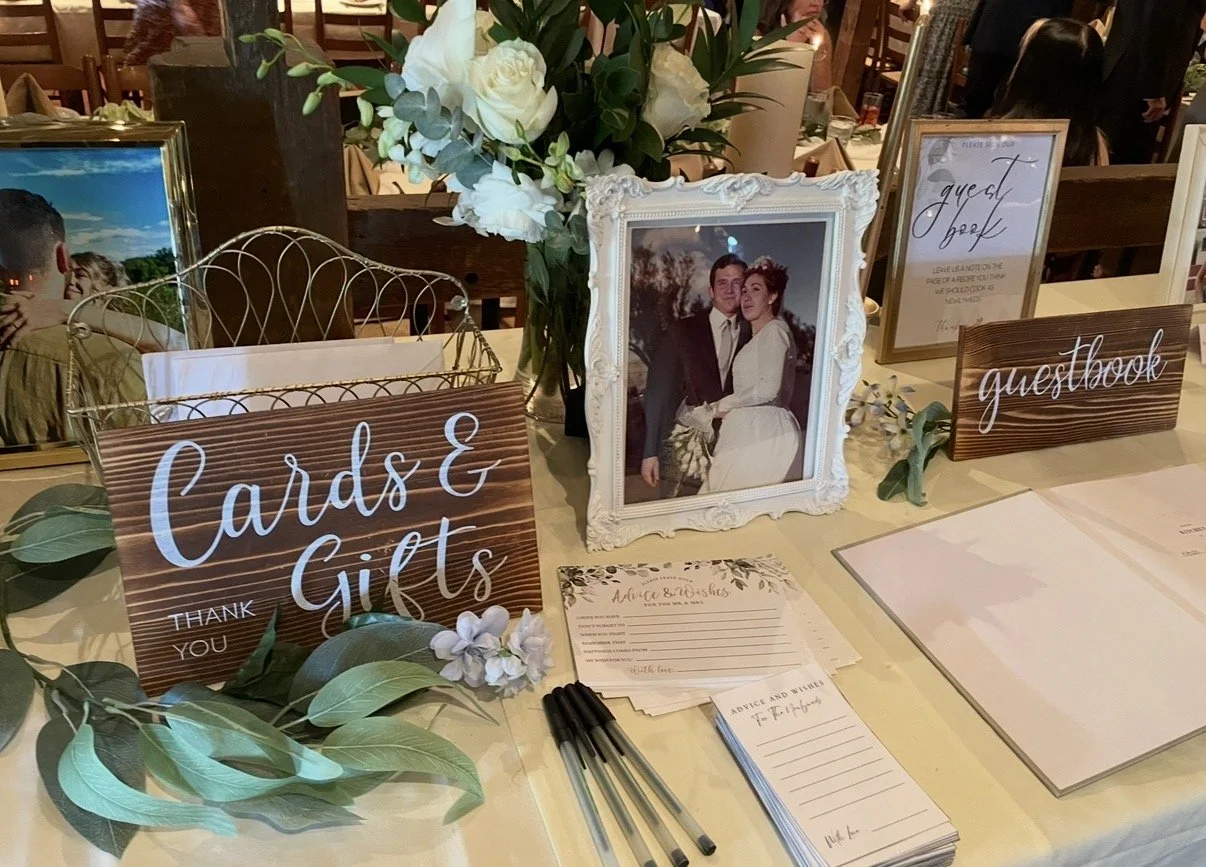 Wedding guestbook and signage table with framed wedding photo, floral arrangement, pens, and advice cards at a wedding reception.