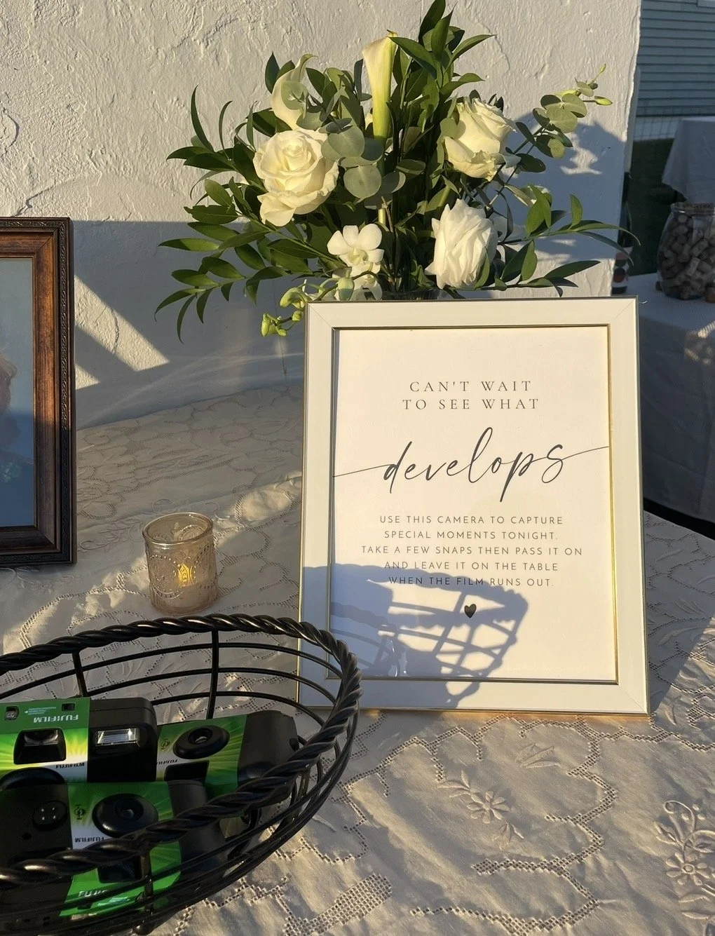 White frame sign with a message about developing and capturing special moments, placed on a lace tablecloth with a bouquet of white roses and greenery behind it. A candle in a glass holder is on the table, and part of a basket with camera batteries and batteries is in the foreground.