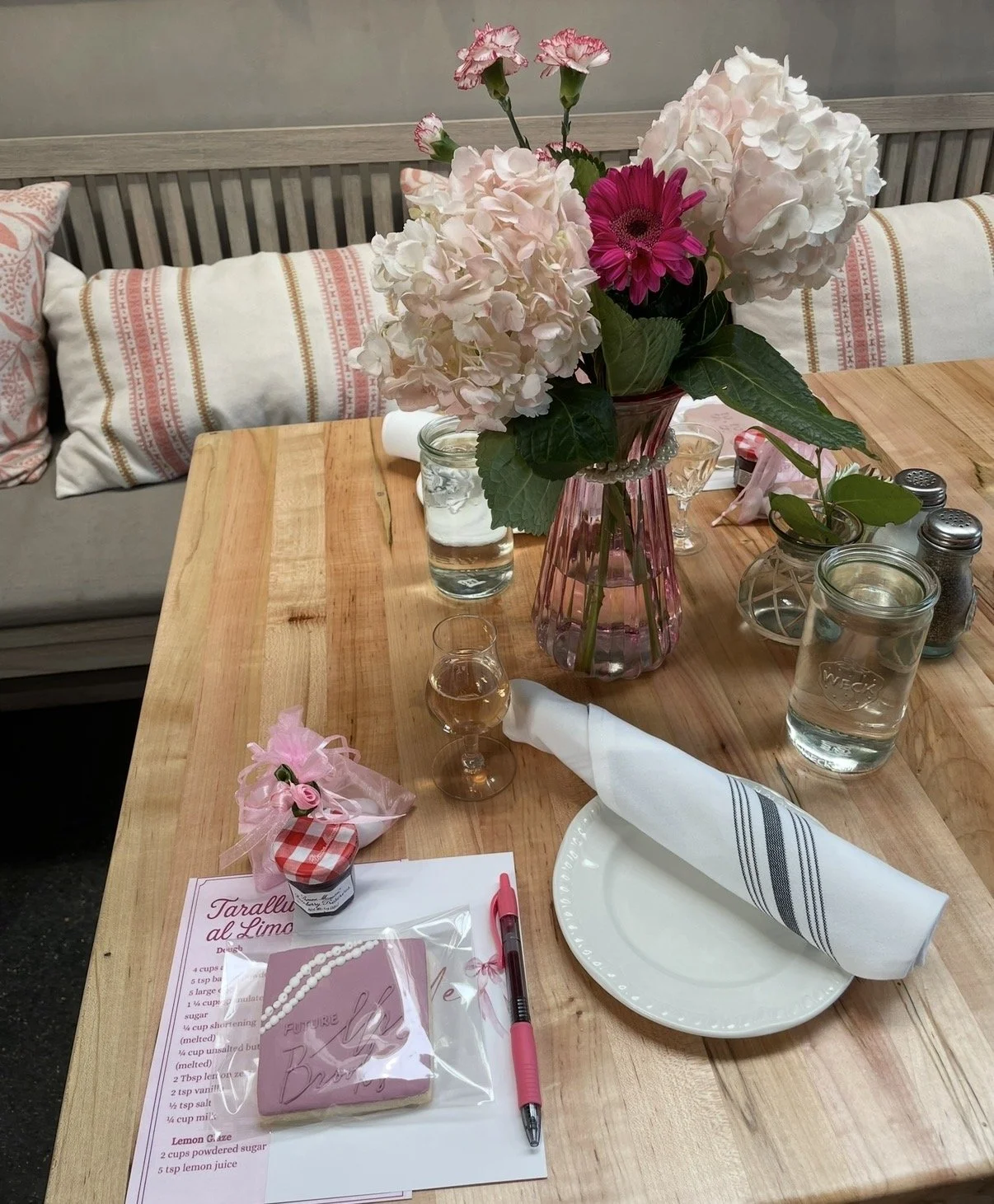 A wooden table decorated with a large pink flower bouquet in a pink vase, glass jars of water, a wrapped pink gift with a pink pen, a plate with a rolled napkin, and small jars of jam or honey, set for a special occasion or brunch.