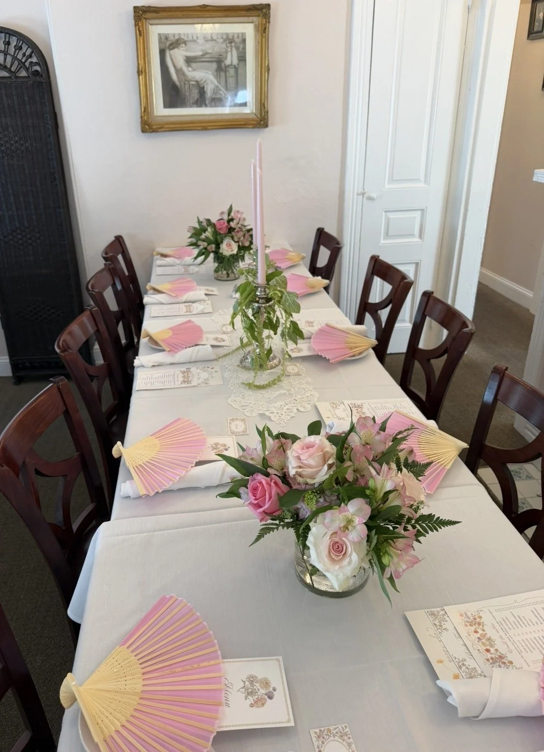 A decorated dining table set for a celebration with pink and white floral arrangements, pink and yellow paper fans, and printed menus or place cards.