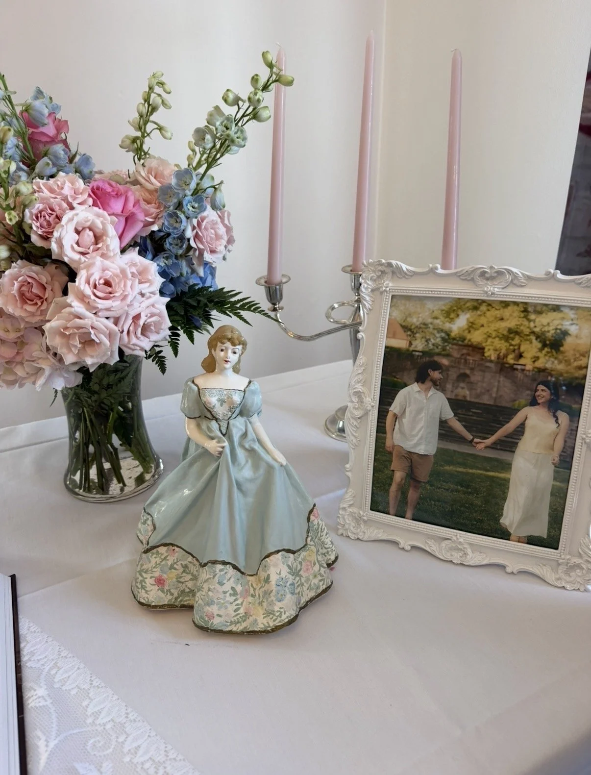 A table decorated with a pink floral bouquet, a porcelain figurine of a woman in a blue dress, a framed photo of a couple holding hands outdoors, and a pink candlestick holding three unlit pink candles.