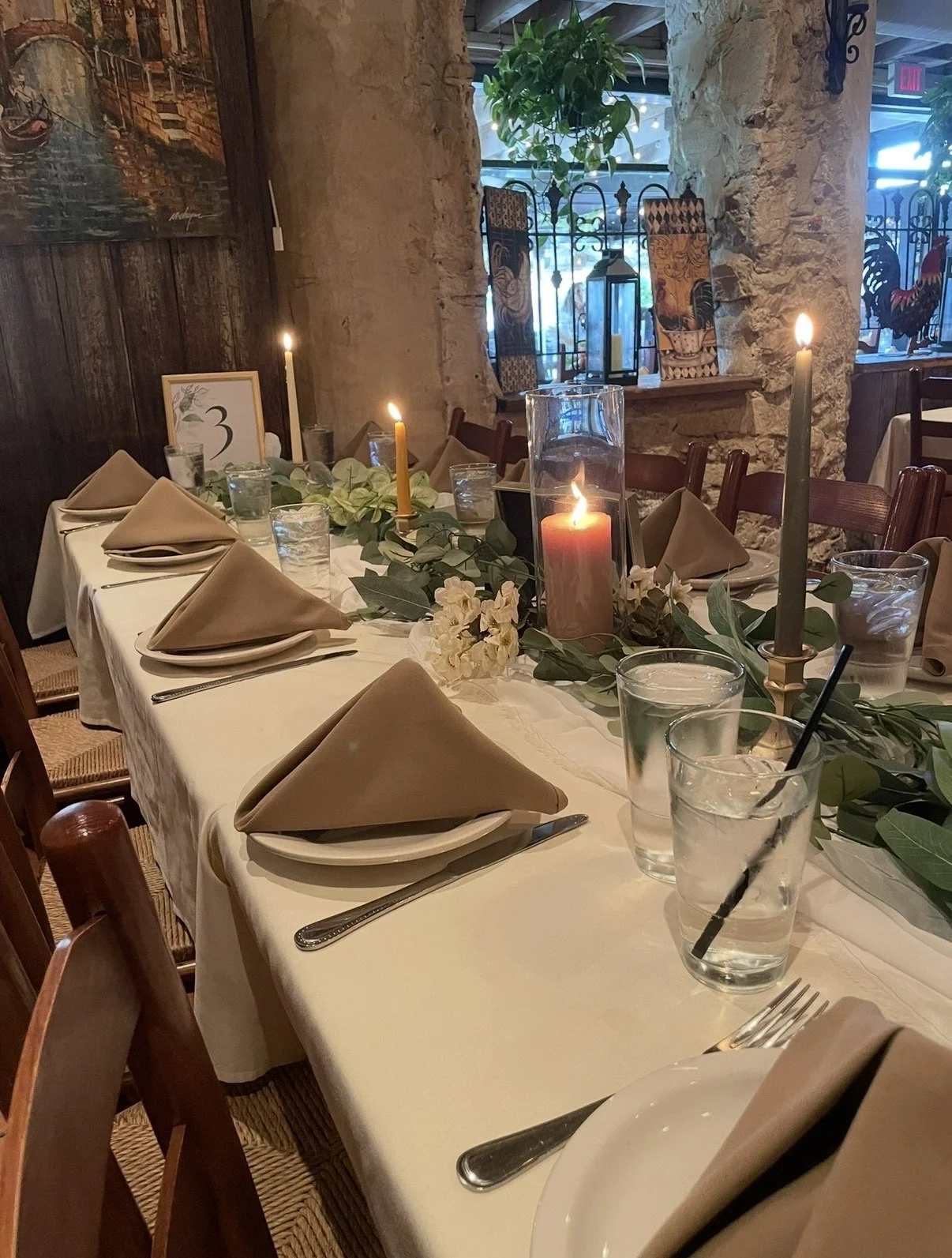 An elegantly set dining table in a restaurant with a white tablecloth, beige napkins folded into triangles, and various glasses and silverware. The table features candles and floral decorations with greenery, and a rustic background with stone walls and artwork.
