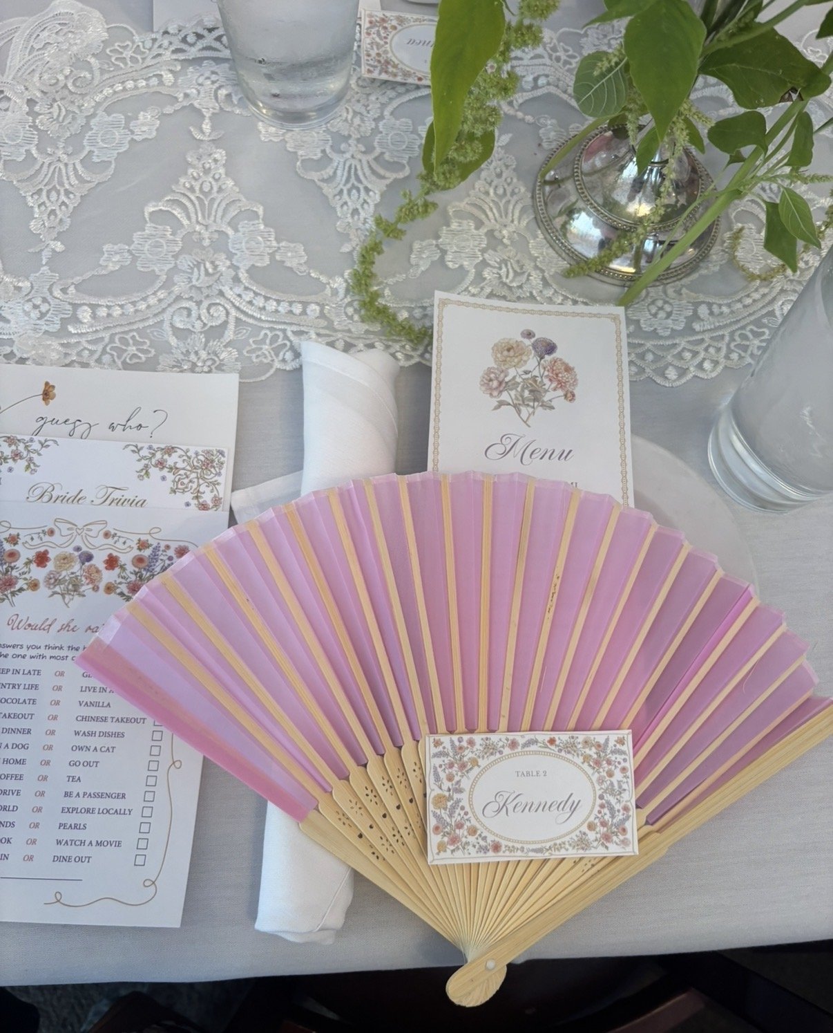 A table setting with a lace tablecloth, a pink and purple folding fan labeled 'Kennedy', menu cards, a glass of water, and a silver vase with green leaves and flowers.
