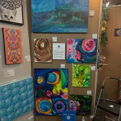 Gallery wall with various colorful abstract paintings and a framed photograph of a gray cat at the top left.