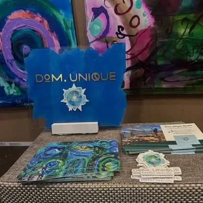 Display table with OM.UNIQUE promotional materials, including a blue sign with the logo, brochures, and business cards, set against colorful abstract paintings.