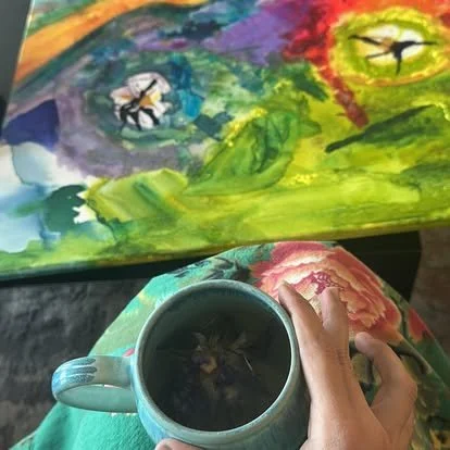 A person holding a mug filled with a dark beverage, with a colorful abstract painting in the background featuring bees and a bird.