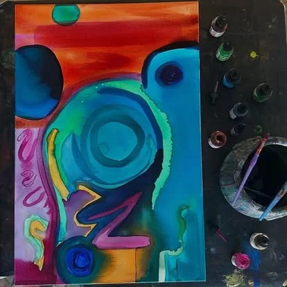 Abstract colorful painting with swirling shapes and vibrant hues, surrounded by bottles of paint and brushes.