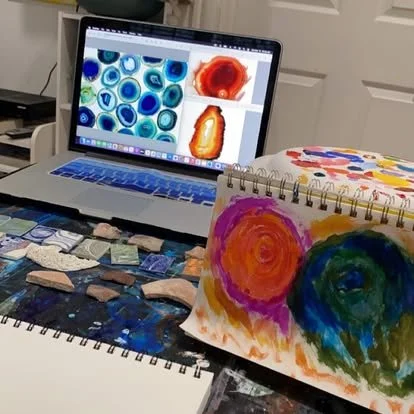A laptop displaying colorful abstract art images on its screen, surrounded by sketchbooks with watercolor paintings, display rocks, and art supplies on a table.