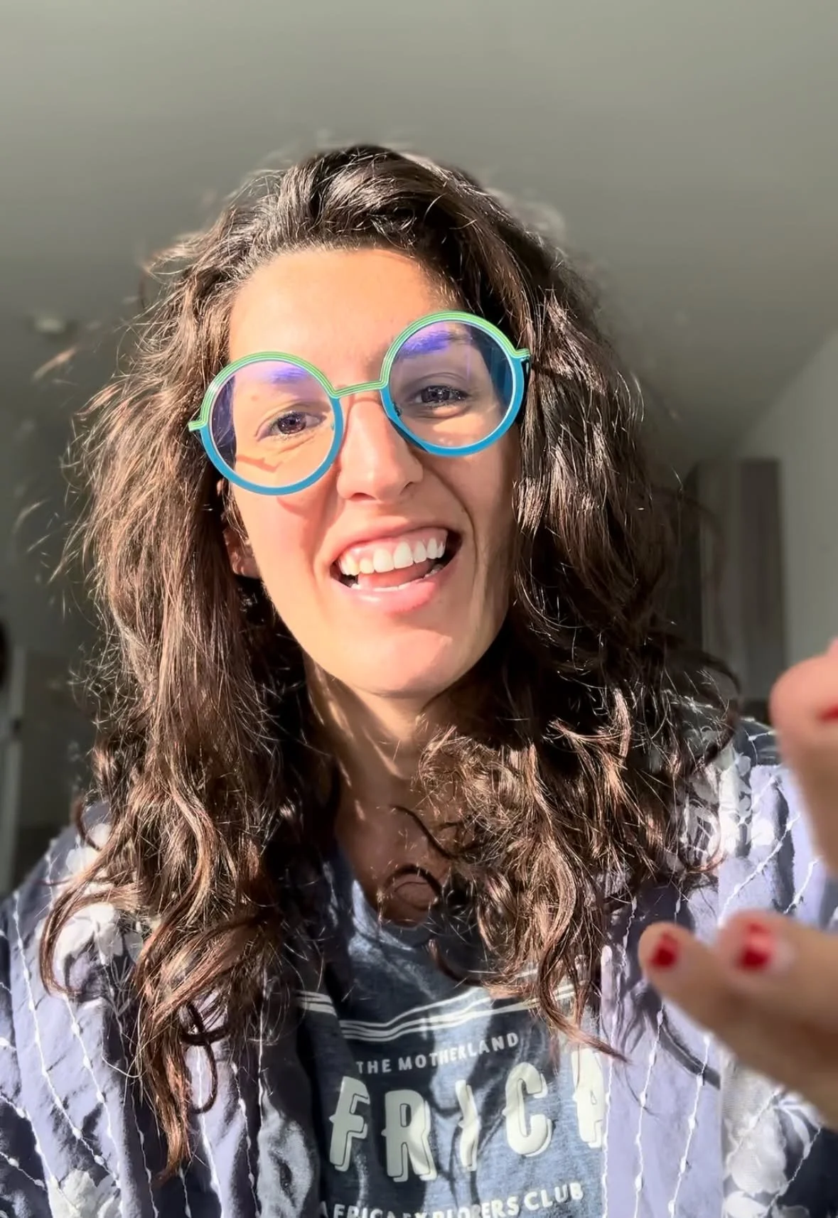 A woman with curly brown hair wearing colorful round glasses smiling at the camera indoor.