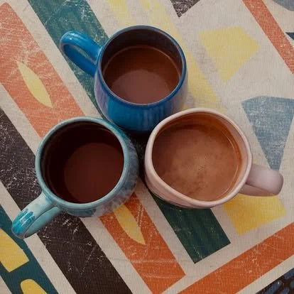 Three colorful coffee mugs filled with hot chocolate on a patterned tablecloth.