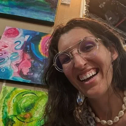 Woman with glasses smiling in front of colorful paintings at an art exhibit.