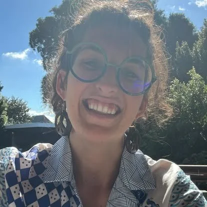 Smiling woman with curly hair and glasses outdoors on a sunny day.