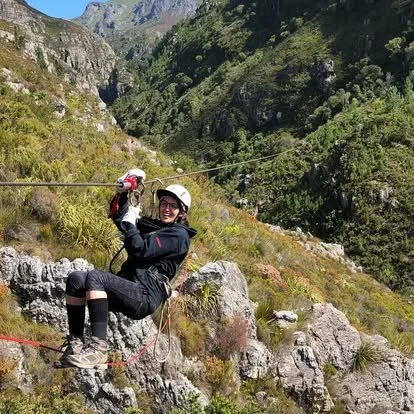 Person in outdoor gear hanging from a zipline over rocky, green mountain landscape.