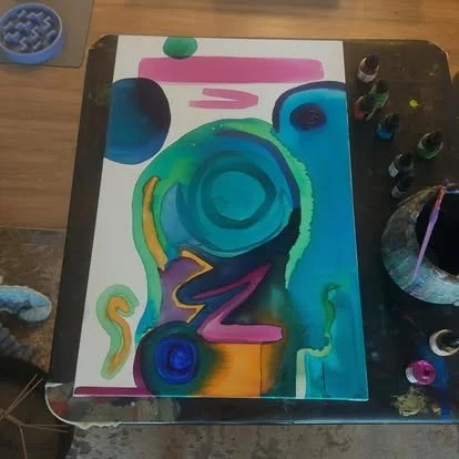 Colorful abstract painting on paper with various shades of blue, green, purple, yellow, and pink, placed on a black table with paint bottles around it.