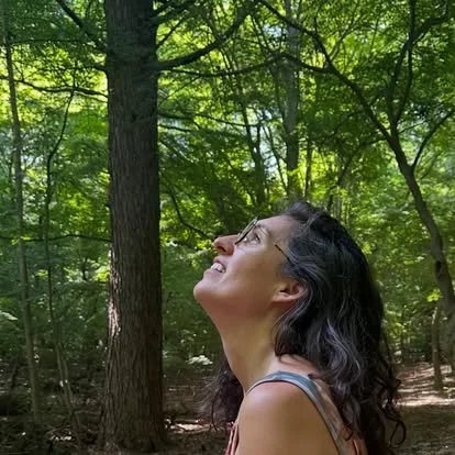 A woman with dark, wavy hair and glasses looking up at the green leafy trees in a forest.