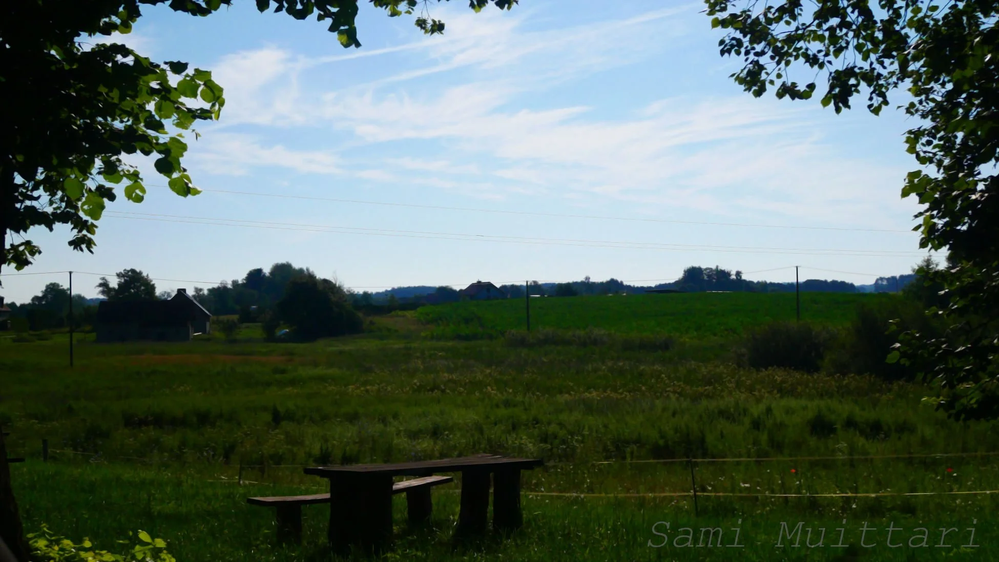 Scenic view of a rural landscape with trees framing the image, green fields, a couple of barn structures in the distance, power lines, and a blue sky with some clouds.