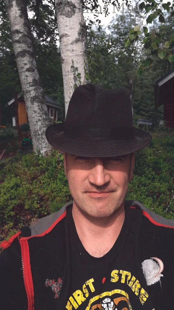 A man wearing a black hat and black and red jacket, smiling outdoors in front of birch trees with green foliage.