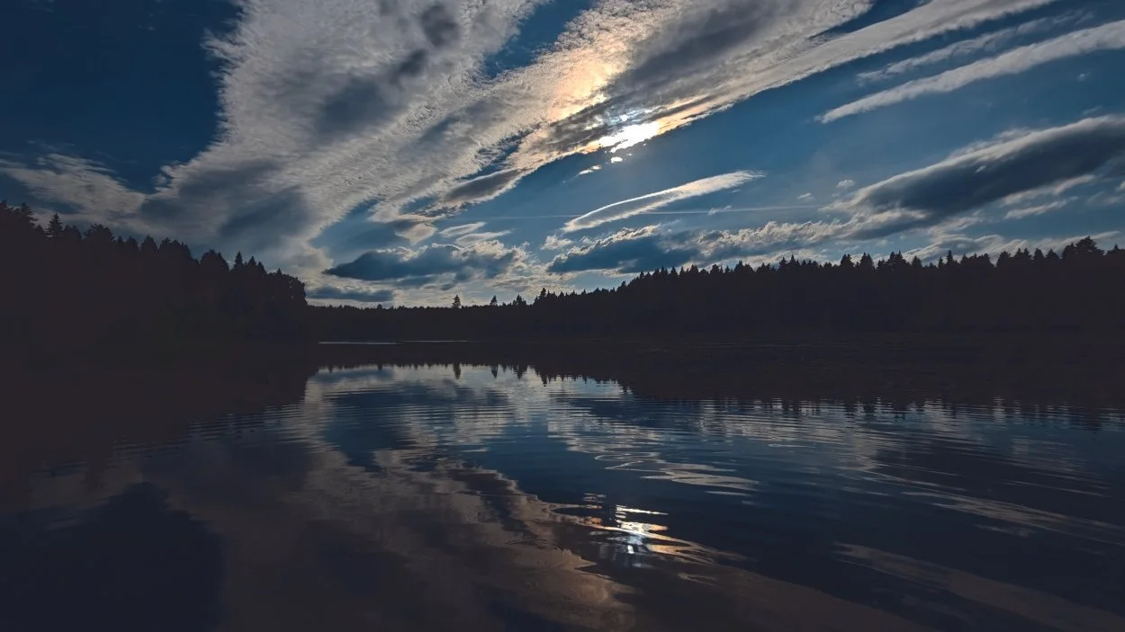 Calm lake reflecting a partly cloudy sky during sunset or sunrise, with silhouetted trees along the horizon.