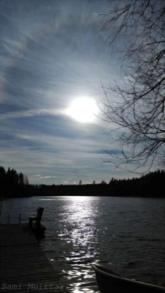 A body of water reflecting sunlight on a clear day with partly cloudy skies, leafless trees on the right, and a wooden dock in the foreground.