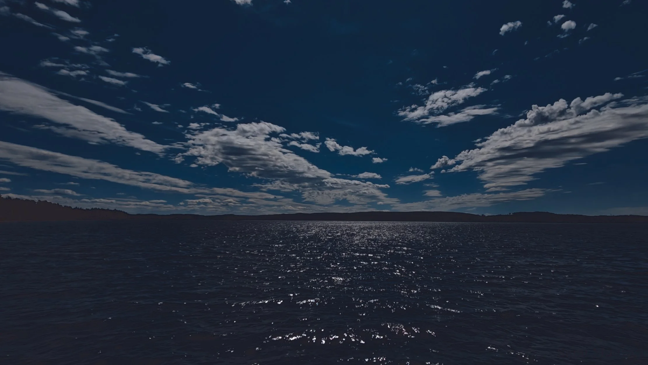 View of a body of water with sunlight reflecting on the surface, under a blue sky with scattered clouds and distant shoreline on the horizon.