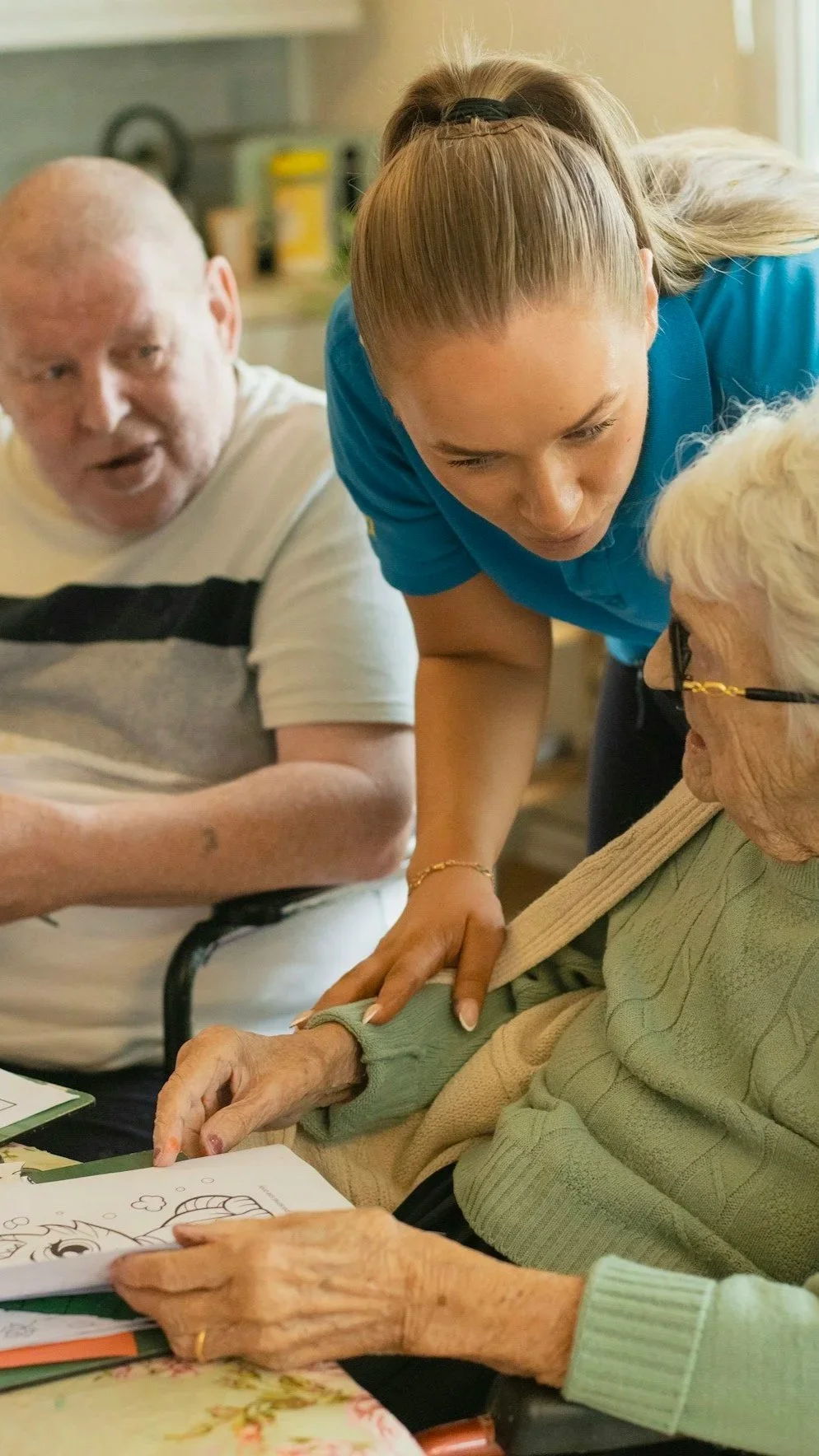 Young woman in blue assisting elderly women with coloring activity at a table.