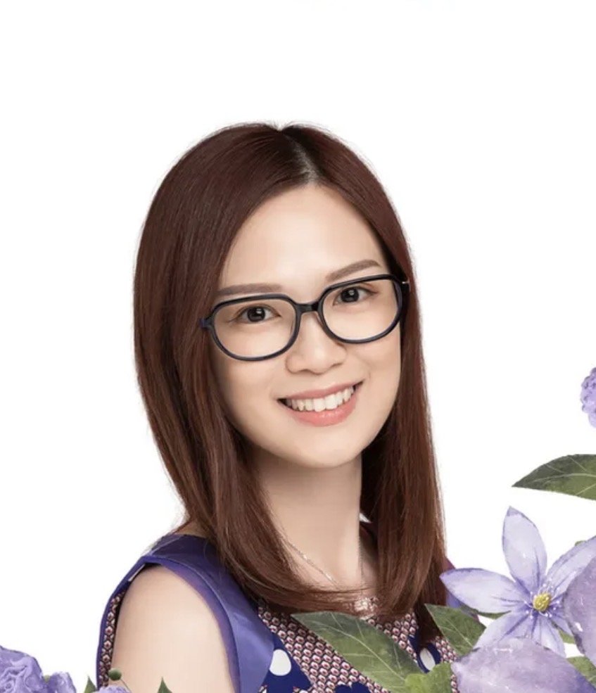 A young woman with shoulder-length brown hair, glasses, and a bright smile, wearing a sleeveless purple dress, surrounded by purple flowers with green leaves, against a white background.