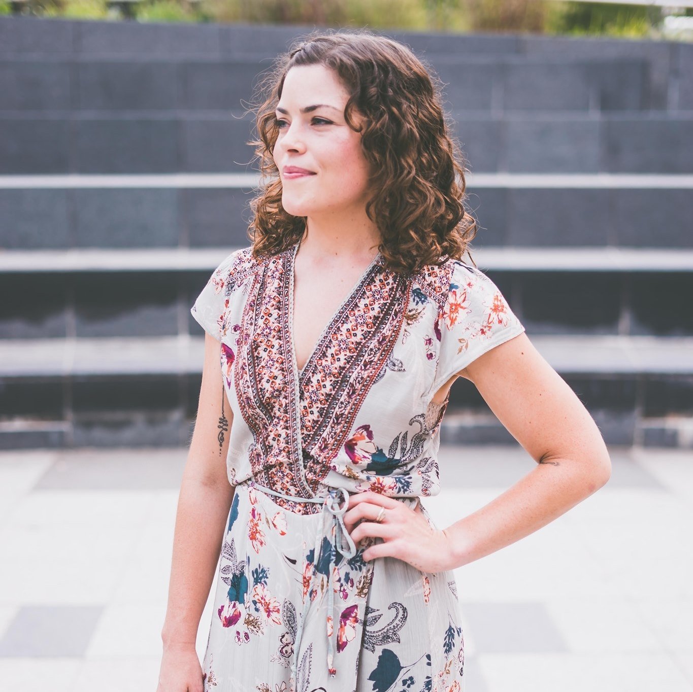 A woman with curly brown hair standing outdoors with her hand on her hip, wearing a short-sleeved, v-neck, floral dress with a waist tie, in front of a staircase or tiered structure.
