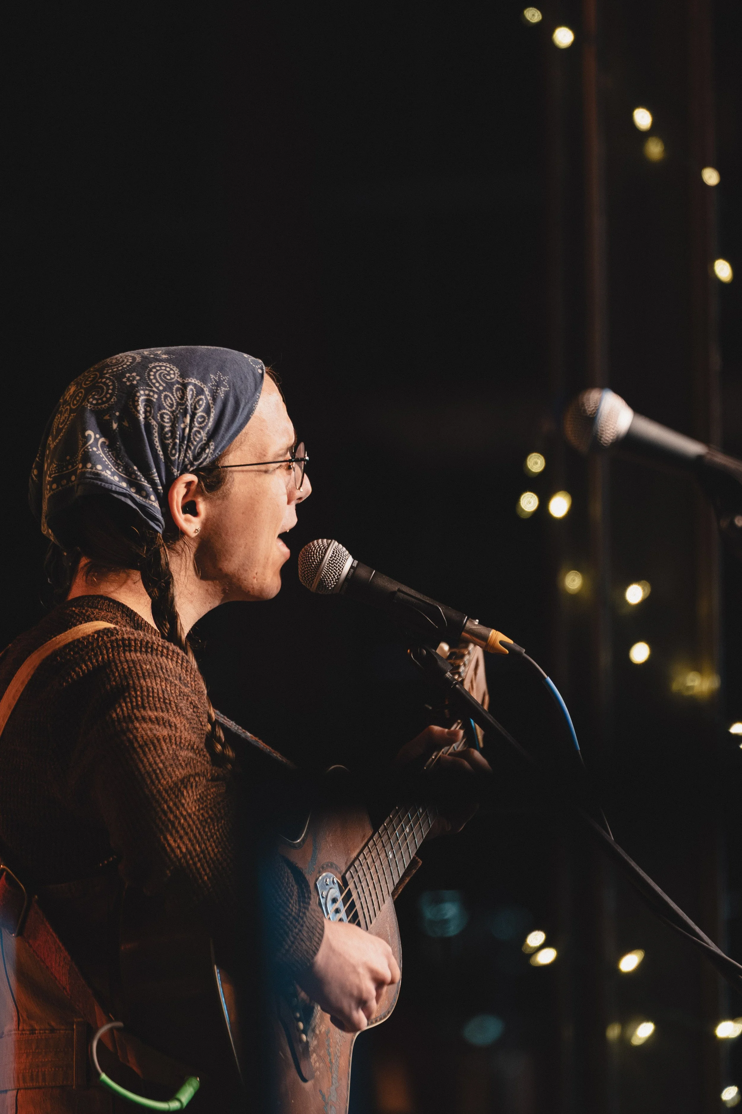 A person wearing glasses, a bandana, and a brown jacket is singing into a microphone while playing a guitar. The background is dark with blurry lights.