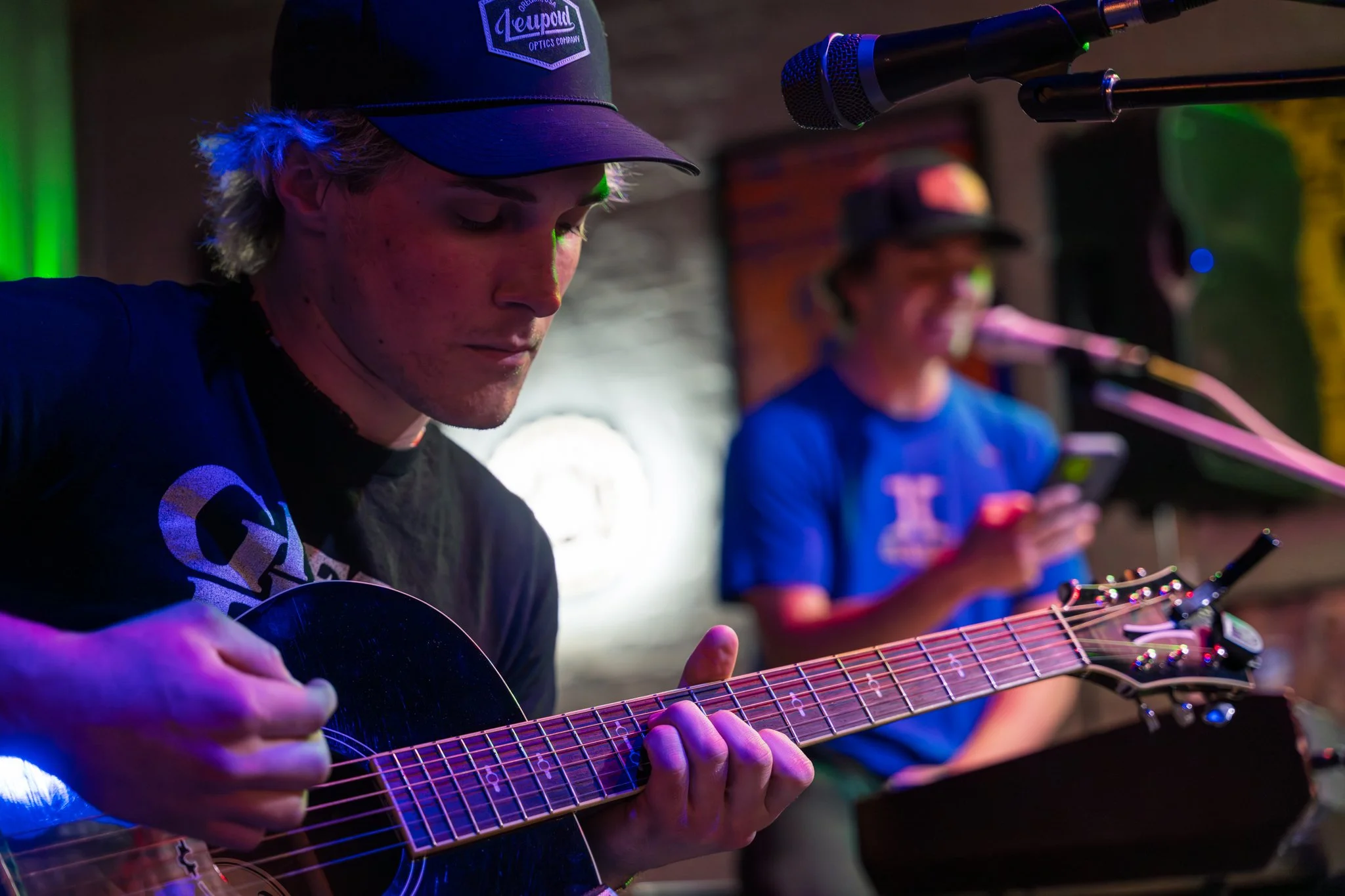 Two young men are playing acoustic guitars and singing into microphones. They are in a dimly lit room with colorful lighting. The man in the foreground wears a black baseball cap with a logo and a black shirt, while the man in the background wears a cap and a blue shirt.