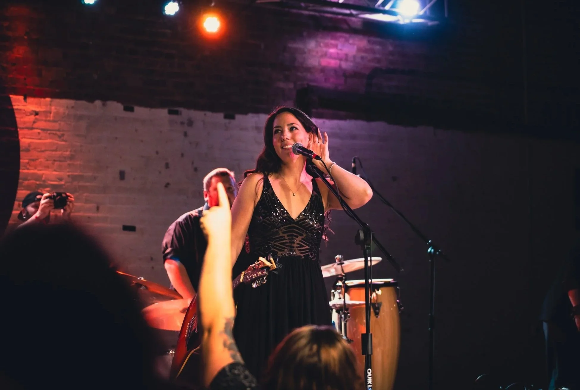 A woman singing on stage with a microphone in hand, wearing a black dress. There are other musicians and instruments, including drums, in the background. Audience members' silhouettes and a person taking photos are visible in the foreground. The setting appears to be a live music venue with brick walls and stage lighting.