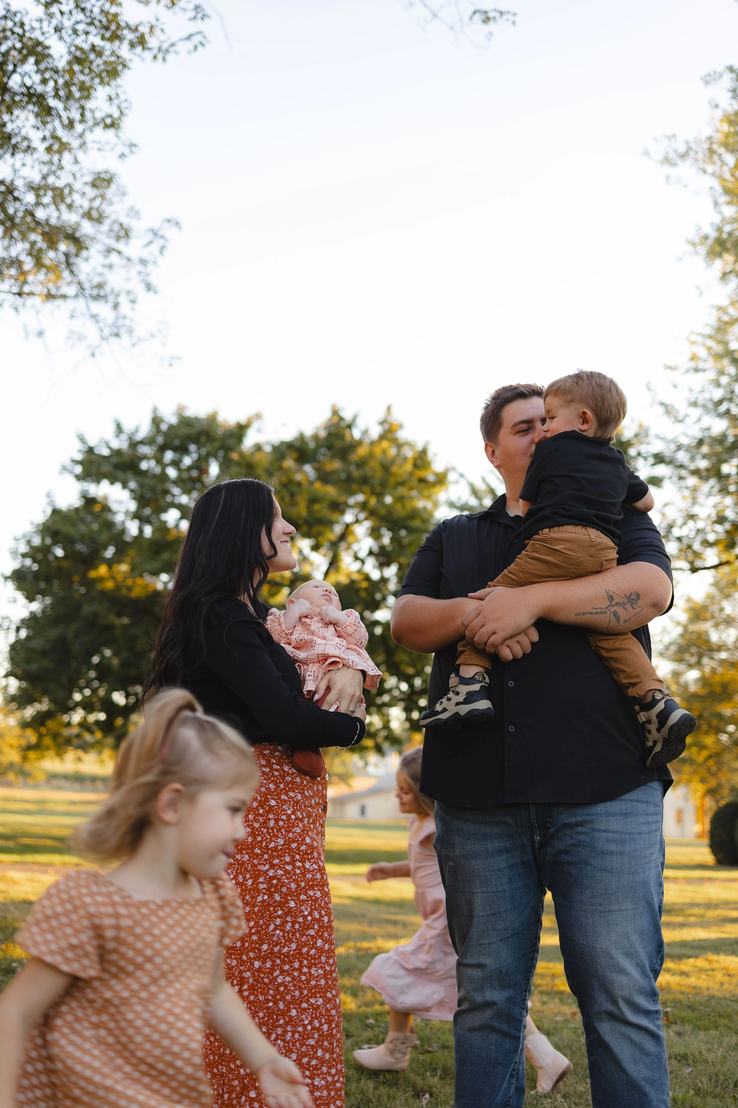 A group of people outdoors in a park during daytime, including a man holding a young child, a woman holding a baby, and children playing.