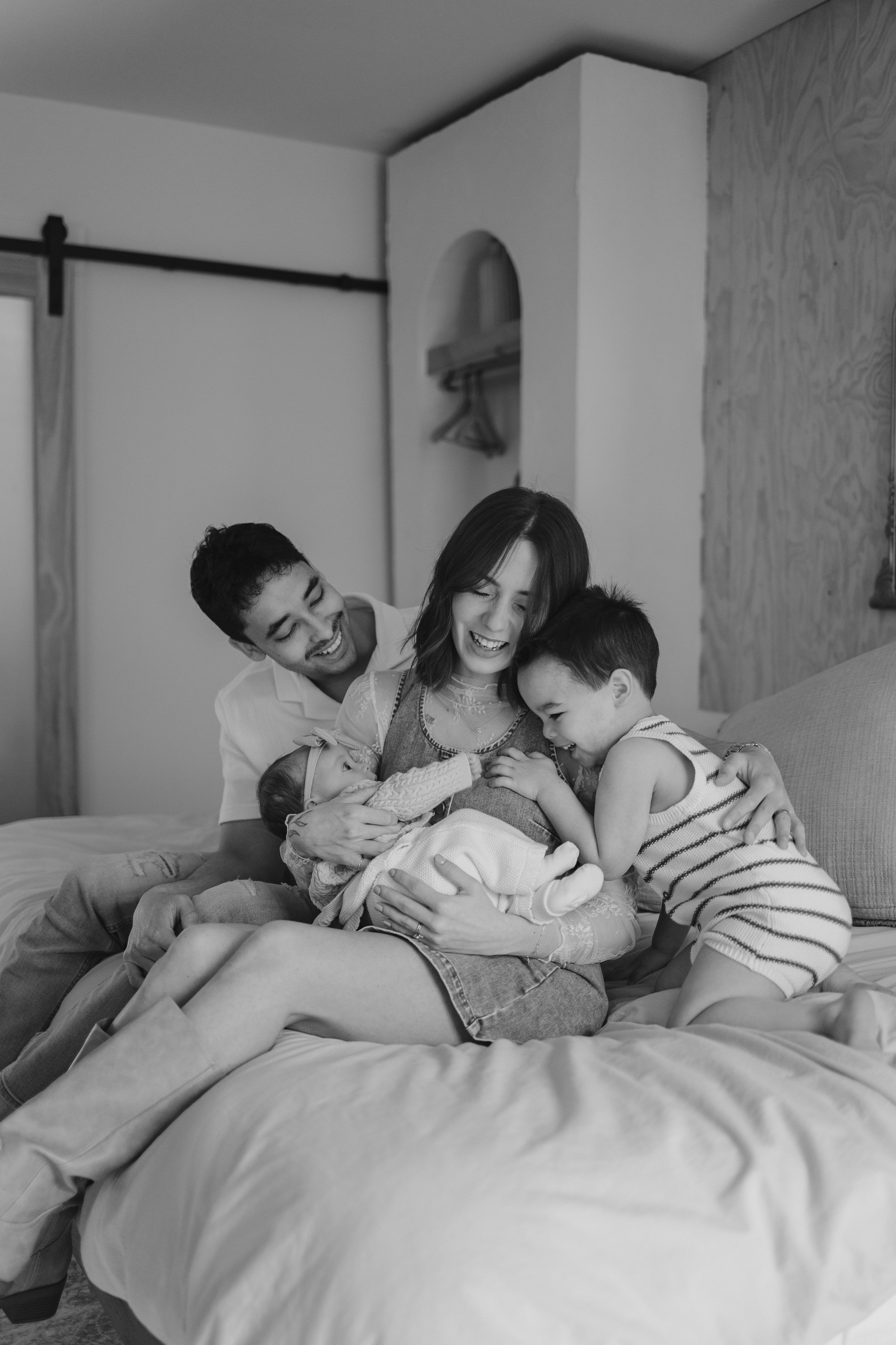 A family of five gathering on a bed, with a woman holding a newborn, a young girl hugging the woman, a young boy leaning on the woman's shoulder, and a man smiling behind them, in a cozy bedroom.