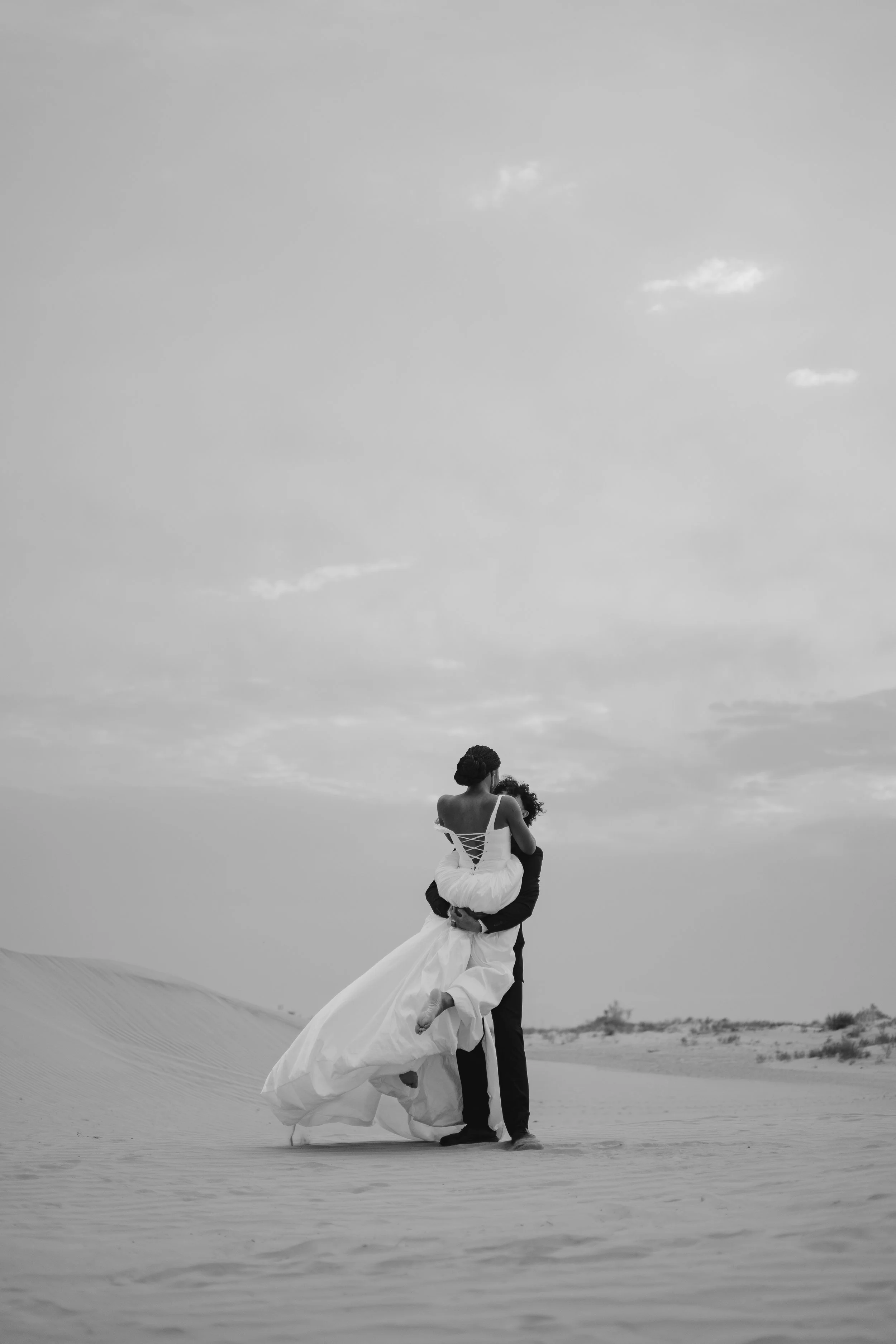 A couple standing in a desert landscape, with the man lifting the woman. The woman is wearing a long white dress, and the man is dressed in black. The scene is in black and white, with a cloudy sky in the background.