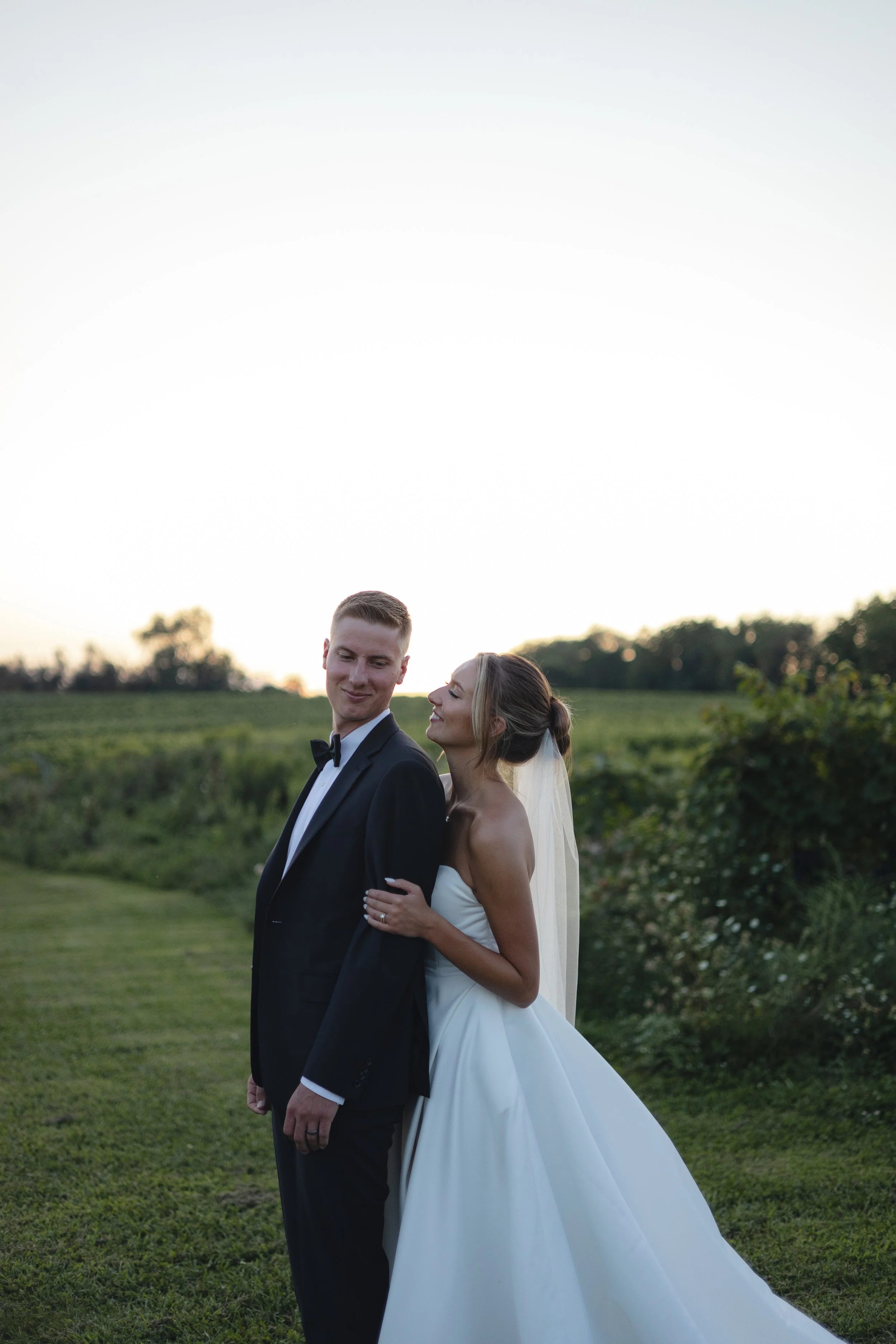 A bride and groom standing outdoors on a grassy field at sunset. The groom wears a black tuxedo with a bow tie, and the bride wears a strapless white wedding gown with a long veil. They are close together, with the bride gently holding the groom's arm, both looking affectionately at each other.