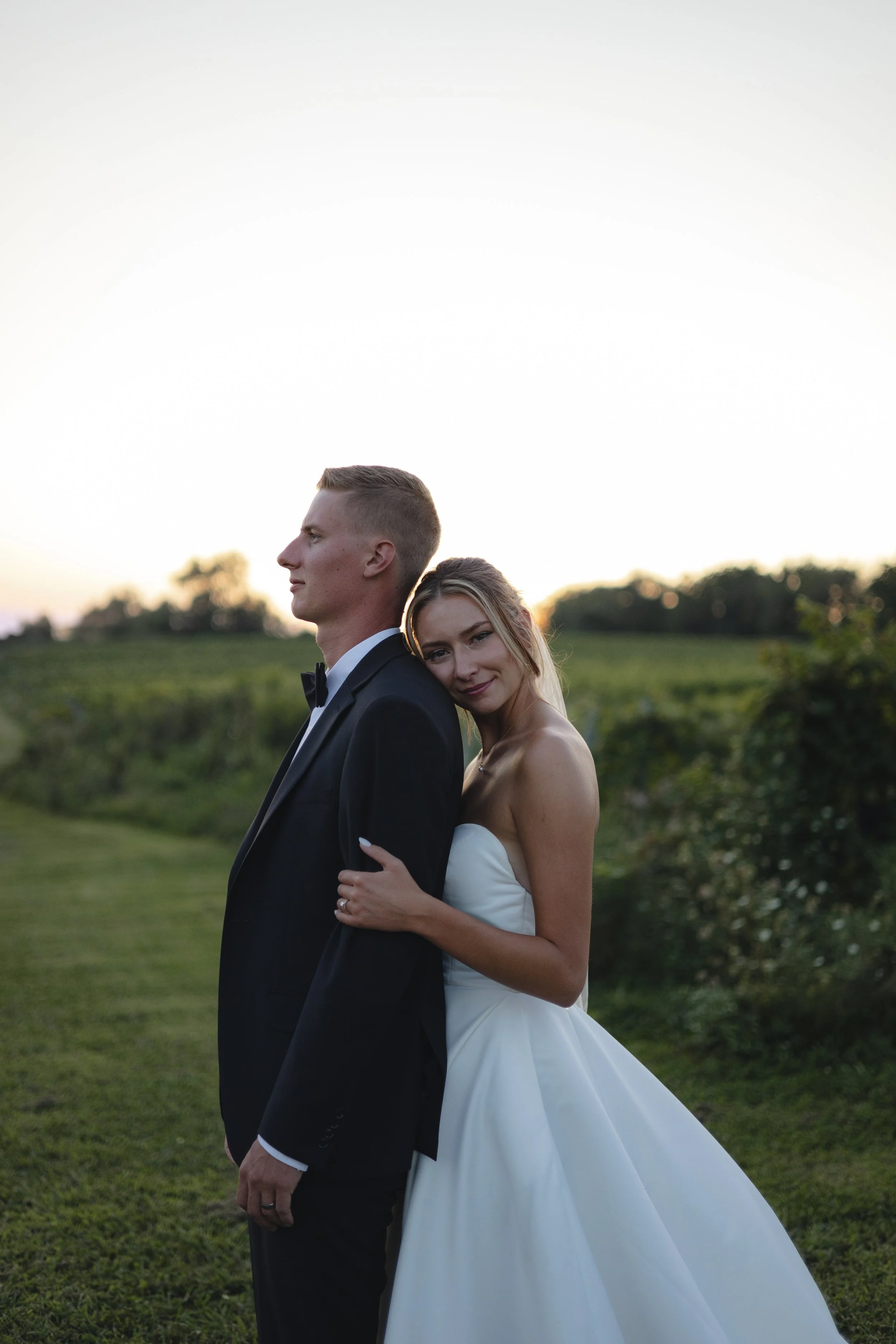 A newlywed couple standing outdoors during sunset; the groom is dressed in a black tuxedo and the bride in a strapless white wedding gown, embracing in a field with trees in the background.