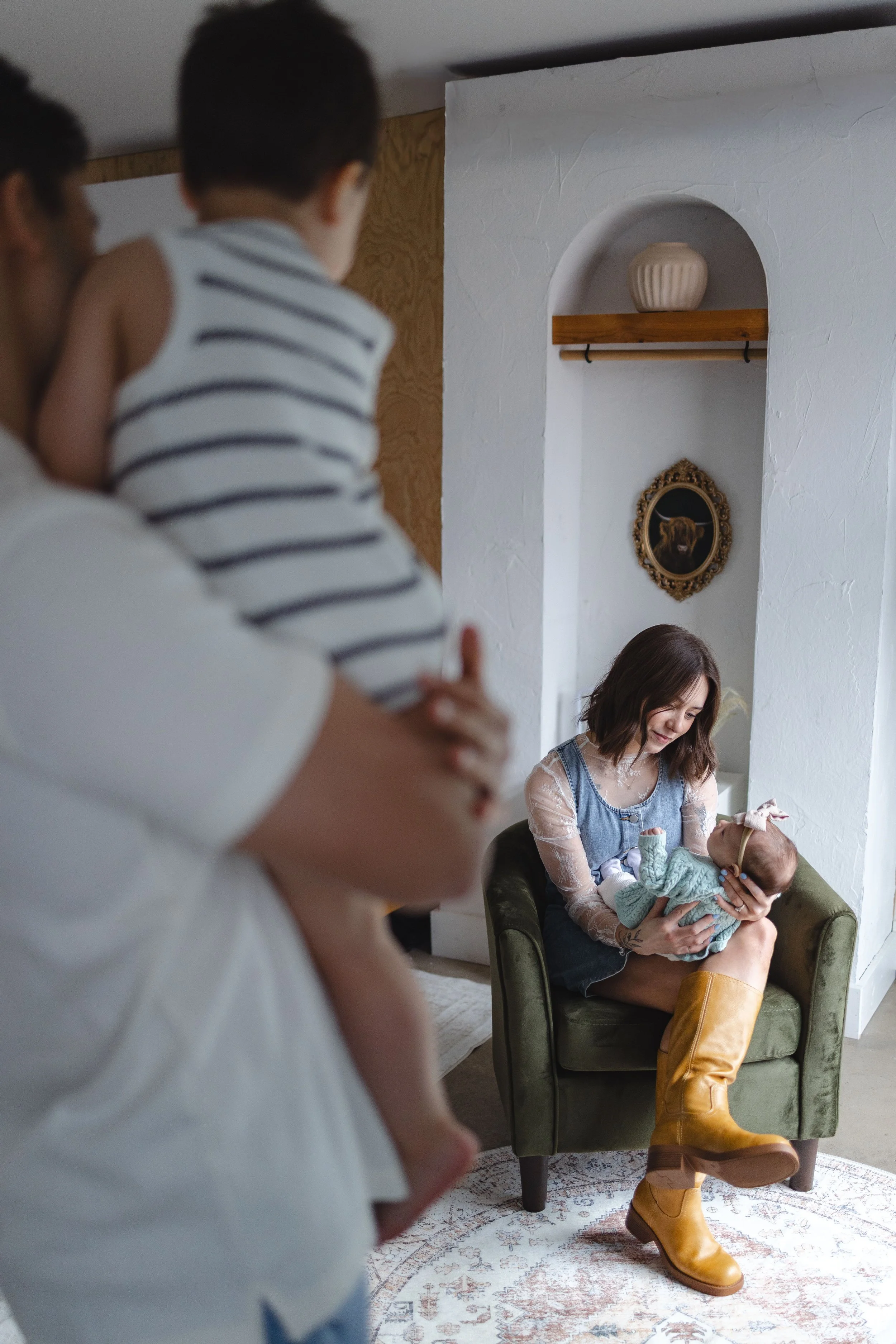 A woman sitting in a green armchair holding a baby, her head inclined downward, smiling, with a young girl beside her, in an indoor setting with wall decor and a patterned rug.