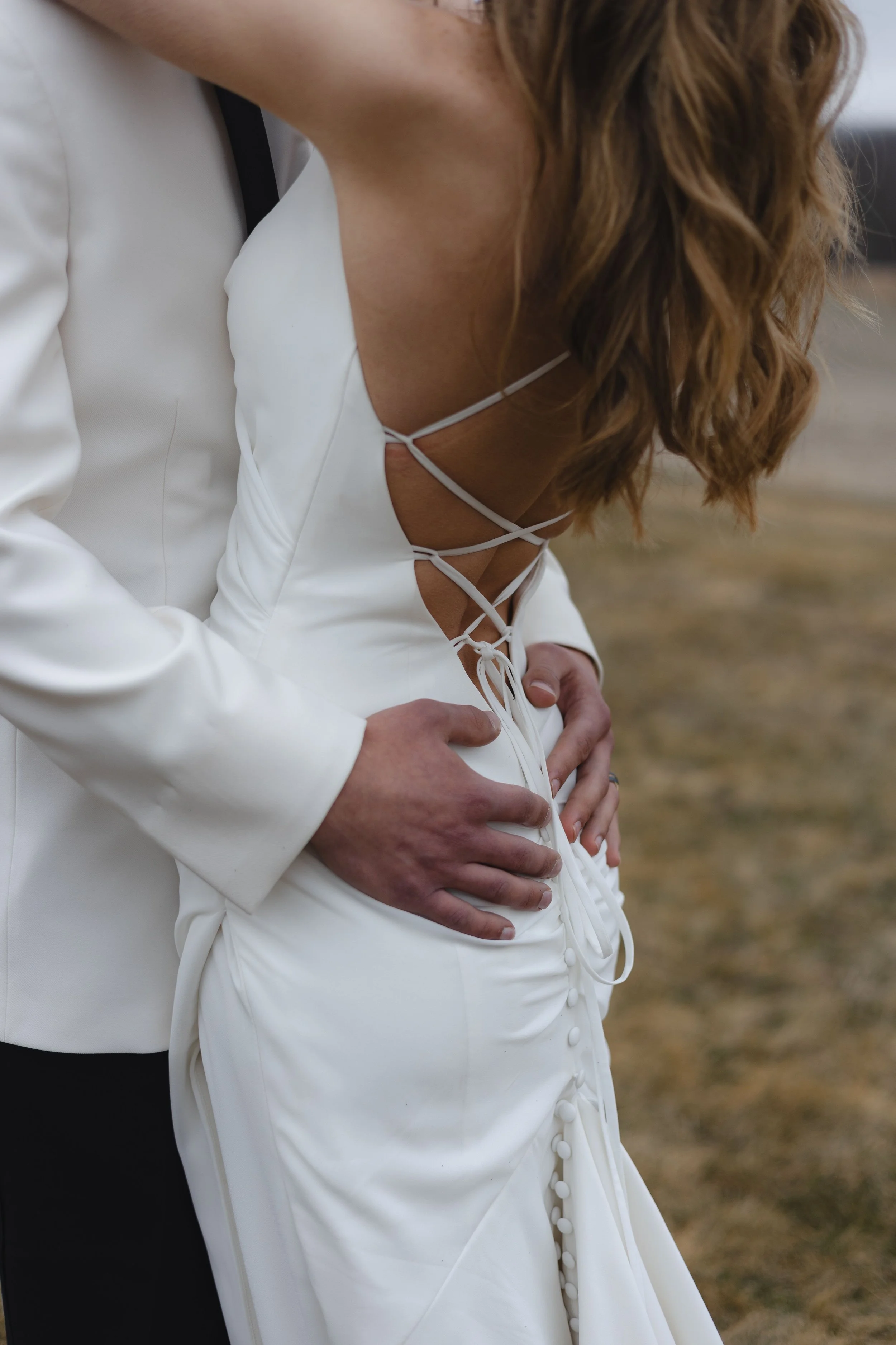 A woman in a white wedding dress is holding her stomach, likely pregnant, with her hands resting on her belly. She has long, wavy brown hair. A man in a suit is standing behind her, partially visible.