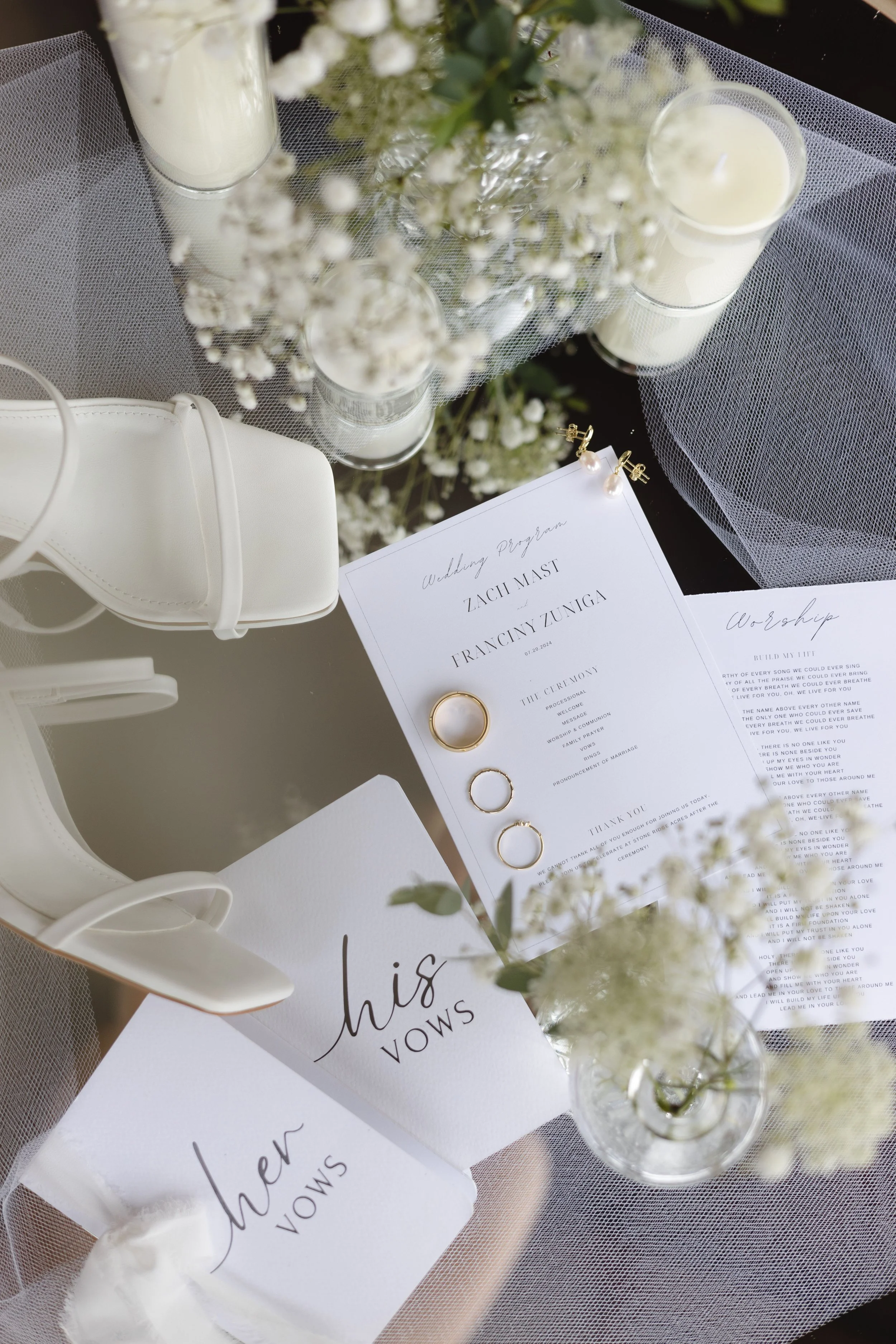 Wedding program, white candles, white floral arrangements in glass vases, rings on a paper, and a white purse, all on a black and white table setting.